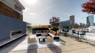 This expansive rooftop terrace is thoughtfully designed to serve as a versatile outdoor amenity space, offering a variety of activity and relaxation zones across its wide, open-air footprint. The surface is finished with large-format concrete pavers, providing a clean, contemporary aesthetic throughout.

A dedicated lounge area anchors one section of the terrace, centered around a sectional sofa and accent chairs arranged on a light-colored area rug. A low-profile coffee table completes the seating arrangement, while a sleek, wall-mounted linear fireplace nearby adds a sophisticated focal point and warmth for cooler evenings. Warm-toned throw pillows accent the neutral upholstery.

Adjacent to the lounge, a games zone features a full-size foosball table set on artificial turf, adding a casual, social element to the space. A hanging egg chair nearby provides a more intimate seating option. Large planters throughout the terrace are filled with ornamental grasses, flowering plants, and mature trees, contributing vibrant color and a sense of natural enclosure.

A bar-height dining area with chairs accommodates smaller gatherings, while glass railings along the perimeter ensure unobstructed panoramic views of the surrounding urban skyline. The rooftop is accessed via a glass-paneled door framed by a wood-accented exterior facade and additional greenery, creating an inviting entry point.