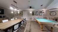 The lounge at 435 Mix is a well-appointed common area designed to serve as both a social and recreational hub for residents. The space is thoughtfully divided into two functional zones. On one side, a full-sized pool table with an ornate wooden base and teal felt surface takes center stage, illuminated by an industrial-style pendant light that adds a refined touch. A cue rack nearby ensures the area is fully equipped for gameplay. On the opposite side, a large rectangular wooden table surrounded by modern white chairs provides an ideal setting for communal dining, meetings, or casual gatherings. This area is lit by a matching set of pendant fixtures, creating a cohesive aesthetic throughout the room. Neutral-toned walls are adorned with framed artwork, contributing to the polished yet relaxed ambiance. Wall sconces offer warm supplemental lighting, and a patterned carpet grounds the space with subtle visual interest. A few additional chairs along the walls offer flexible seating. The overall design balances functionality and comfort, making this lounge a welcoming destination for socializing and leisure activities.