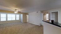 This single-floor residence features an open-concept layout that connects the living and dining areas, creating a spacious and functional flow. The living area is finished with wall-to-wall carpet in a neutral beige tone, contributing to a warm and understated aesthetic throughout. A ceiling fan with integrated lighting is centrally mounted, providing both air circulation and ambient illumination. Neutral-toned walls complement the light-colored baseboards and trim, lending the space a clean, cohesive appearance. A window along the far wall allows natural light to filter into the living area, enhancing the sense of openness. A hallway visible in the background suggests access to additional rooms, likely including a bedroom and bathroom. The kitchen is partially visible to the right, separated by a raised countertop bar with dark laminate surfaces and white cabinetry, offering a practical divide between the cooking and living spaces. HVAC vents are integrated into the walls and ceiling, ensuring climate control throughout. Overall, the space presents a straightforward, move-in-ready layout suited for everyday living.