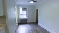 This single-floor unit presents a straightforward and functional layout with classic architectural details throughout. The main living area is finished with rich, dark hardwood floors that add warmth and character to the space. Crown molding runs along the ceiling perimeter, lending a traditional touch to the otherwise neutral, light-toned walls. A ceiling fan with an integrated light fixture is centrally mounted, providing both ventilation and ambient lighting. Natural light enters through a standard window fitted with a wall-mounted air conditioning unit, offering year-round climate control. A cast-iron radiator beneath the window provides supplemental heating, consistent with classic urban residential construction. The room connects to additional spaces through two separate doorways, suggesting a multi-room layout with clear circulation between areas. The white-painted door and window trim throughout provides clean contrast against the soft gray-white walls. The overall aesthetic is simple and unfurnished, offering a blank canvas that can be adapted to a variety of uses. The combination of traditional building elements — hardwood floors, crown molding, and radiator heating — alongside modern conveniences creates a practical and timeless living environment.