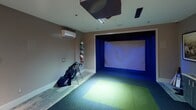 Located at 4307 Sharon Rd in Charlotte, NC, this single-floor sports entertainment space is thoughtfully designed around a professional-grade indoor golf simulator. The simulator bay anchors the room, featuring a large projection screen framed by blue accent lighting, a synthetic turf hitting mat, and an overhead projector mounted to the ceiling — creating an immersive, stadium-like atmosphere for year-round play. A dedicated wall-mounted mini-split system ensures climate comfort throughout sessions.

Adjacent to the simulator bay, a built-in cabinetry station serves as the tech and control hub, housing a monitor that displays simulator software, a beverage refrigerator, and ample surface space for scorecards and accessories. A framed vintage sports photograph adds a classic, clubhouse-inspired character to the wall above. A vertical wall-mounted ball display showcases a curated collection of sports balls, adding a playful, athletic aesthetic to the space.

The lounge area offers two round bistro-height tables paired with upholstered bar chairs finished in warm taupe and chrome, providing comfortable seating for spectators and guests. Framed golf patent blueprints decorate the walls, reinforcing the sporting theme. Hardwood-style flooring transitions seamlessly into the carpeted simulator zone, and recessed ceiling lighting throughout creates an intimate, polished ambiance suited for both casual recreation and competitive play.