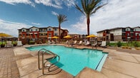 Located at 7430 N 100th Ave in Glendale, Arizona, this outdoor amenity space offers a well-appointed resort-style retreat designed for relaxation and recreation in the heart of the Sonoran Desert. The centerpiece of the space is a generously sized swimming pool with crystal-clear turquoise water, complemented by a separate spa and a shallow wading area, all finished with clean concrete and paver surrounds. Towering palm trees frame the pool deck, providing natural shade and a striking visual backdrop against the Arizona sky. Ample lounge chairs and vibrant yellow umbrellas are arranged throughout the sundeck, offering plentiful spots for sunbathing and leisure. A covered pergola with a metal roof provides a shaded gathering area, alongside dedicated BBQ and grilling stations ideal for outdoor entertaining. Adjacent to the pool, a brick-clad clubhouse or amenity building with large openings connects seamlessly to the outdoor environment. Landscaped areas featuring desert-friendly gravel, native plants, and patches of artificial turf add greenery and texture to the hardscape. Decorative brick paver pathways tie the entire space together, creating a cohesive and inviting atmosphere. The surrounding multi-story residential buildings provide a sense of community while the open sky above enhances the spacious, airy feel of this outdoor amenity area.