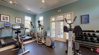 This fitness center offers a thoughtfully designed workout environment that balances functionality with an inviting aesthetic. The space is finished with soft teal walls, crisp white crown molding, and recessed lighting, lending it a clean, polished atmosphere. Rubber flooring throughout provides both durability and comfort underfoot during workouts.

The cardio section is well-stocked with multiple treadmills, elliptical trainers, a stair climber, and stationary and spin bikes, several of which are equipped with personal media screens. A water cooler station is conveniently positioned for easy access. The strength-training area features a multi-station cable machine, a dedicated Ab Coaster, an adjustable bench, and a full rack of dumbbells organized by weight, accommodating a wide range of resistance training routines.

A wall-mounted flat-screen television is positioned for visibility across the space, and a large mirror along one wall visually expands the room while allowing users to monitor their form. Natural light pours in through multiple windows fitted with blinds, and French doors open directly to an outdoor area, providing views of surrounding greenery and allowing for excellent ventilation. A ceiling fan adds to the comfort of the space. The overall ambiance is bright, airy, and purpose-built for a productive fitness experience.