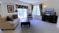 This two-floor residence offers a thoughtfully designed layout that balances comfort and style across its generous living spaces. The main living area is a standout feature, bathed in abundant natural light streaming through multiple large windows dressed with decorative valances and coordinating drapes. The lush greenery visible through the glass creates a seamless connection to the outdoors, enhancing the airy and welcoming atmosphere. A sliding glass door opens directly onto a private balcony, extending the living space and providing a peaceful outdoor retreat. Underfoot, plush wall-to-wall carpeting adds warmth and comfort to the room, which is furnished with a spacious sofa, tufted leather ottoman, and an accent chair with matching footstool — all arranged to encourage relaxed conversation and everyday living. Soft wall sconces complement the natural light, lending a warm ambiance during evening hours. A media console with shelving anchors one corner of the room. The residence spans two floors, offering distinct zones for living, entertaining, and private retreat, making it a versatile and functional home suited to a variety of lifestyles.