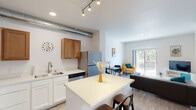 This thoughtfully designed single-floor unit offers a seamless open-concept layout that connects the kitchen, dining, and living areas into one cohesive, light-filled space. The kitchen is well-appointed with warm wood upper cabinetry, white lower cabinets, light quartz countertops, stainless steel appliances including a range, microwave, dishwasher, and refrigerator, and a central island with bar seating. An exposed metal duct adds an industrial design accent overhead, complemented by track lighting. Light wood-style flooring runs throughout the main living areas, adding warmth and continuity. The living area is bright and inviting, featuring a sliding glass door that opens to a private balcony with views of surrounding greenery, allowing ample natural light to fill the space. The bedroom is a serene retreat finished with plush carpet, a large window with outdoor views, and direct access to a dedicated walk-in closet offering generous storage. The bathroom features a quartz-topped vanity, wood-style flooring, and a shower-tub combination. A dedicated laundry room houses a full-size side-by-side washer and dryer with overhead shelving and a hanging rod, providing exceptional in-unit convenience.