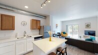 This thoughtfully designed single-floor unit offers a seamless open-concept layout that connects the kitchen, dining, and living areas into one cohesive, light-filled space. The kitchen is well-appointed with warm wood upper cabinetry, white lower cabinets, light quartz countertops, stainless steel appliances including a range, microwave, dishwasher, and refrigerator, and a central island with bar seating. An exposed metal duct adds an industrial design accent overhead, complemented by track lighting. Light wood-style flooring runs throughout the main living areas, adding warmth and continuity. The living area is bright and inviting, featuring a sliding glass door that opens to a private balcony with views of surrounding greenery, allowing ample natural light to fill the space. The bedroom is a serene retreat finished with plush carpet, a large window with outdoor views, and direct access to a dedicated walk-in closet offering generous storage. The bathroom features a quartz-topped vanity, wood-style flooring, and a shower-tub combination. A dedicated laundry room houses a full-size side-by-side washer and dryer with overhead shelving and a hanging rod, providing exceptional in-unit convenience.
