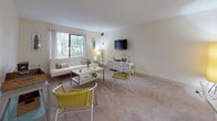 This single-floor residence offers a thoughtfully arranged layout connecting key living areas in a cohesive, functional flow. The living room serves as the heart of the space, featuring plush carpeting, neutral walls, and a large window that frames outdoor greenery, filling the room with natural light. A shelving unit provides additional storage, while the open layout comfortably accommodates multiple seating areas.

Adjacent to the living room, the dining area is defined by a pendant light overhead and a pass-through opening that connects directly to the kitchen, creating an efficient and sociable arrangement. The kitchen is equipped with warm wood cabinetry, light countertops, a coil-burner electric range, and tile flooring, offering ample storage and workspace.

The bedroom is generously sized with soft carpeting, neutral tones, and a large window with vertical blinds that allow for adjustable natural light. A walk-in closet provides notable storage capacity. Bedside lamps on both sides create a balanced, comfortable atmosphere.

The bathroom features a warm wood vanity with a broad countertop, an integrated sink, and tile flooring, maintaining the clean, understated aesthetic found throughout the residence. Overall, the space presents a calm, neutral palette with practical finishes throughout.