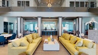 The Tamarac Village clubhouse is a thoughtfully designed community amenity space that blends modern architecture with inviting, upscale interiors. The grand lobby immediately commands attention with soaring double-height ceilings and a wall of floor-to-ceiling windows that flood the space with natural light and offer views of the surrounding landscape. Vibrant yellow sofas, a clean white coffee table, and warm wood-toned flooring establish a welcoming, contemporary atmosphere throughout.

A prominent staircase with rich wood-finish treads and sleek stainless steel railings serves as both a functional and architectural focal point, leading to an upper-level mezzanine overlooking the main floor. A reception desk bearing the Tamarac Village branding anchors the entry area.

The interior transitions seamlessly into a social bar and lounge area, featuring dark cabinetry, backlit shelving, and bar-height seating, ideal for gatherings. Adjacent to this is a well-appointed kitchen and dining zone, highlighted by a substantial granite island with gold-toned barstools and a striking cluster pendant light fixture. Coffered wood-accented ceilings and expansive glazed walls connect this area to the outdoor pool deck beyond.

A separate dining room with a rustic dark wood table and glass doors provides direct outdoor access, completing a space designed for community, comfort, and connection.