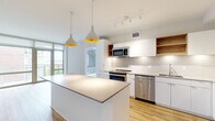 This contemporary single-floor residence showcases a clean, modern design throughout. The open-concept kitchen is a standout feature, outfitted with flat-front white cabinetry, warm wood open shelving, a tiled backsplash, and a full suite of stainless steel appliances including a refrigerator, range, built-in microwave, and dishwasher. A large central island with a light quartz countertop provides additional workspace and seating, while stylish pendant lights with gold interiors add a warm, designer touch overhead. Track lighting further illuminates the space.

The living area features light hardwood-style flooring and is anchored by expansive floor-to-ceiling sliding glass doors that open to a balcony, flooding the room with natural light and offering outdoor views. The bedrooms are finished with plush carpeting and include closet access, maintaining a calm and comfortable atmosphere.

The bathrooms are thoughtfully designed with large-format tile flooring, floating white vanities, and quartz countertops with undermount sinks. A full-width mirror enhances the sense of space. One bathroom also conveniently houses an in-unit washer and dryer behind louvered doors. Throughout the residence, a cohesive neutral palette of whites, grays, and natural wood tones creates a bright, contemporary ambiance.
