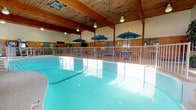 This single-story indoor pool facility is designed to provide a comfortable and functional aquatic recreational experience year-round. The centerpiece of the space is a generously sized freeform swimming pool with vivid turquoise water, complemented by a separate hot tub nearby. A safety railing encloses the pool perimeter, and life rings are mounted along the walls for added security.

The architecture features warm natural wood paneling along the lower walls and striking exposed wood beams supporting a vaulted ceiling, creating a rustic yet inviting ambiance. A continuous row of large windows lines one side of the facility, flooding the poolside with natural light and offering views of the surrounding outdoor landscape.

The lounge area adjacent to the pool is furnished with blue strap chairs and high-top tables topped with teal patio umbrellas, ideal for relaxing between swims. The entry area includes vending machines, restroom access, a water fountain, and informational bulletin boards. Pool rules and safety signage are clearly posted throughout, including a designated chemical storage area. The durable terrazzo-style flooring throughout the facility is practical and easy to maintain. The space is well-ventilated with integrated HVAC vents, ensuring a comfortable environment for all visitors.