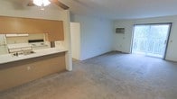 This single-floor residential space is designed with an open, functional layout that maximizes the available living area. The main living space features wall-to-wall carpeting in a neutral tone, contributing to a warm and comfortable atmosphere. A ceiling fan with a light fixture is centrally positioned overhead, providing both ventilation and ambient lighting throughout the room.

The kitchen is partially separated from the living area by a half-wall partition, creating a pass-through counter that maintains an open feel while defining each space. The kitchen is equipped with white appliances, including a range and microwave, set against light-colored cabinetry and countertops. A cork board or wooden accent panel adds a practical touch to the kitchen area.

A wall-mounted air conditioning unit is installed along one of the exterior walls, ensuring climate control throughout the space. Natural light enters the room through a sliding glass door located along the far wall, which also provides direct access to an outdoor area, enhancing the sense of openness and connectivity to the exterior. The overall color palette is light and neutral, with white walls that keep the space feeling bright and airy throughout.