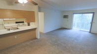This single-floor residential space is designed with an open, functional layout that maximizes the available living area. The main living space features wall-to-wall carpeting in a neutral tone, contributing to a warm and comfortable atmosphere. A ceiling fan with a light fixture is centrally positioned overhead, providing both ventilation and ambient lighting throughout the room.

The kitchen is partially separated from the living area by a half-wall partition, creating a pass-through counter that maintains an open feel while defining each space. The kitchen is equipped with white appliances, including a range and microwave, set against light-colored cabinetry and countertops. A cork board or wooden accent panel adds a practical touch to the kitchen area.

A wall-mounted air conditioning unit is installed along one of the exterior walls, ensuring climate control throughout the space. Natural light enters the room through a sliding glass door located along the far wall, which also provides direct access to an outdoor area, enhancing the sense of openness and connectivity to the exterior. The overall color palette is light and neutral, with white walls that keep the space feeling bright and airy throughout.