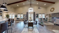 This clubhouse is defined by its dramatic vaulted ceiling with exposed wooden beams, creating an open and airy atmosphere throughout the main gathering space. Large windows and glass French doors flood the interior with natural light, offering views of the surrounding outdoor areas. The great room serves as the heart of the clubhouse, anchored by a central round dining table beneath a statement geometric chandelier, with distinct lounge seating areas arranged on either side. One lounge area highlights a striking floor-to-ceiling herringbone tile fireplace surround with a modern linear electric fireplace, complemented by neutral sofas and teal accent décor. Large-format tile flooring in a cool grey tone runs throughout, unifying the space. The reception area near the entrance features decorative columns, a tray ceiling with accent lighting, and a staffed front desk alongside digital kiosks for resident convenience. The well-appointed kitchen includes crisp white shaker cabinetry, granite countertops, a mosaic tile backsplash, stainless steel appliances, and a breakfast bar with pendant lighting. The overall ambiance is sophisticated yet inviting, blending contemporary design elements with warm, transitional styling throughout.