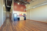This single-floor urban loft showcases an industrial-chic aesthetic defined by soaring ceilings with exposed structural beams, open ductwork, and overhead track lighting. Rich, dark hardwood flooring runs throughout the main living areas, lending warmth and contrast to the raw architectural elements above.

The open-concept living space is generously proportioned, offering flexibility in layout and design. Large windows along one wall flood the space with natural light, highlighting the exposed brick that adds texture and character to the interior.

The kitchen is efficiently designed along a single wall, featuring light-toned cabinetry, a full suite of black appliances including a refrigerator, electric range, built-in microwave, and dishwasher, as well as a double-basin sink with a bold, earthy accent wall backdrop.

The bedroom area transitions to plush carpeting and offers an abundance of storage with multiple sliding-door closets, making excellent use of the expansive wall space. The high, exposed-beam ceiling continues into this room, maintaining the loft's cohesive industrial character.

Two full bathrooms provide practical functionality, each featuring a tub-shower combination, toilet, and tile flooring. One bathroom includes a vanity with under-cabinet storage and a large mirror, adding a refined touch to the utilitarian design.