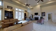 This single-floor communal living space features an open, airy layout highlighted by impressively tall ceilings with a tray ceiling detail and dual ceiling fans that contribute to a sense of grandeur. Large glass-paneled doors with transom windows along one wall flood the interior with natural light while offering views of the surrounding outdoor greenery. The light-toned tile flooring runs throughout, enhancing the bright and welcoming atmosphere. A traditional wood-burning fireplace with a classic white mantle serves as a central focal point, topped by a wall-mounted television and flanked by a framed artwork piece. Seating is generously arranged throughout, including plush sofas and upholstered armchairs in neutral and muted tones, accommodating multiple conversation areas. A built-in bookcase with display shelving adds a functional yet decorative element near the entrance area. Recessed lighting and natural light work together to create a warm, inviting ambiance. The space is accented with area rugs, potted plants, and tasteful decor, making it a comfortable and well-appointed gathering area suitable for relaxing or socializing.