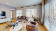 This well-appointed single-floor residence combines contemporary design with thoughtful details across its living spaces. The open-concept kitchen is outfitted with light wood cabinetry, granite countertops, stainless steel appliances including a gas range and built-in microwave, and a spacious island with seating — illuminated by elegant pendant lighting. The kitchen flows naturally into the living and dining areas, where large windows flood the space with natural light and offer views of the surrounding urban streetscape. The living room features light hardwood flooring, recessed lighting, and a generous layout accommodating sophisticated furnishings. The dining area is anchored by a marble-top table beside floor-to-ceiling windows. The primary bedroom is distinguished by textured accent walls, wall sconces, and large windows with drape treatments. A second bedroom offers a bright, airy feel with ample natural light. The primary bathroom features a dual-sink vanity with wood cabinetry set against rich, deep-toned walls. The second bathroom includes a glass-enclosed walk-in shower, a single vanity, and tile flooring. A central hallway connects all rooms, decorated with framed artwork and finished with warm hardwood flooring throughout.