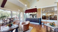 This well-appointed clubhouse in Jacksonville, Florida offers a thoughtfully designed single-story gathering space that balances comfort with functionality. The main lounge area immediately captures attention with its dramatic vaulted ceilings, pendant lighting, and rich hardwood flooring that flows throughout the space. A prominent service bar, accented by a bold burgundy overhead canopy and dark cabinetry, serves as the focal point of the main area and includes a coffee station and water dispenser, making it an ideal hub for residents and guests alike. Comfortable lounge seating — including plush barrel chairs and a deep leather sofa arranged around a stone-topped coffee table — creates an inviting atmosphere for casual gathering and relaxation. Large floor-to-ceiling windows and glass doors flood the interior with natural light while offering picturesque views of lush, mature Florida landscaping and the outdoor patio area beyond. A dedicated business center features a wall-length desk lined with computer workstations set against expansive windows, providing both a productive workspace and a scenic backdrop of the surrounding greenery. Warm, earthy tones on the walls complement the wood floors throughout, giving the entire clubhouse a cohesive, welcoming ambiance suited for both social and professional use.