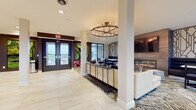 This single-floor amenity lounge is thoughtfully designed to blend contemporary aesthetics with functional communal spaces. The entry area makes a striking first impression, featuring large glass double doors flanked by two lush living moss wall installations set against dark-painted accent walls. Light-toned large-format tile flooring flows throughout, providing a clean, cohesive foundation across the entire space.

A circular candelabra-style chandelier serves as a central design focal point, casting warm ambient light over the open floor plan. Structural columns are integrated seamlessly into the layout, helping define distinct zones without enclosing the space. A dedicated lounge area offers deep navy upholstered booth seating arranged around a central fireplace unit, above which a wall-mounted television is installed. Geometric laser-cut wall panels frame the lounge area, adding an artful, architectural dimension.

A fully equipped kitchenette features sleek white cabinetry, dark stone countertops, a stainless steel sink, and a three-dimensional sculptural tile backsplash. Open shelving displays decorative accents, and an adjacent dining area with floor-to-ceiling windows offers views of the surrounding exterior. Near the main entrance, a secondary seating nook with rounded accent chairs provides a welcoming reception area, completing this well-appointed communal space.