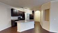 This single-story space showcases an open-concept layout that seamlessly connects the kitchen and living areas, creating a functional and cohesive flow. The kitchen is a standout feature, equipped with rich, dark espresso cabinetry that provides ample storage alongside a suite of black appliances, including a range, microwave, and refrigerator. A central kitchen island with a granite countertop and an integrated sink serves as both a practical workspace and a natural gathering point. Crown molding runs along the upper edges of the walls, adding a refined architectural detail that elevates the overall aesthetic. Track lighting illuminates the kitchen workspace effectively, while the warm-toned walls complement the dark cabinetry throughout. Rich, dark hardwood-style flooring extends across the space, grounding the design with a consistent and polished finish. A graceful arched entryway leads toward what appears to be the front entrance, with a paneled door visible in the background, lending a classic architectural touch. The open sightlines between the kitchen and adjacent living area enhance the sense of spaciousness, making the layout feel welcoming and well-suited for both everyday living and entertaining.