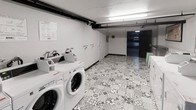 This single-floor laundry facility is designed for shared use and offers a practical, well-organized layout. The room accommodates multiple front-loading washing machines lined along one wall, each equipped with individual detergent dispensers mounted above for convenience. A row of dryers is also visible toward the far end of the space, providing a complete wash-and-dry setup in one location.

The flooring features a distinctive geometric patterned tile in grey and white tones, adding a decorative touch to an otherwise utilitarian environment. The walls are finished in plain white, keeping the space bright and clean in appearance. Overhead, fluorescent strip lighting runs along the ceiling, ensuring the room is well-lit throughout.

Exposed pipework runs along the upper portion of the wall, a characteristic detail that reflects the functional, no-frills nature of the facility. An instructional notice is posted on the wall near the machines, likely providing usage guidelines for occupants. A doorway at the far end leads further into the building, suggesting this laundry room is part of a larger shared facility. Overall, the space is compact yet efficiently arranged to serve its purpose.