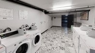 This single-floor laundry facility is designed for shared use and offers a practical, well-organized layout. The room accommodates multiple front-loading washing machines lined along one wall, each equipped with individual detergent dispensers mounted above for convenience. A row of dryers is also visible toward the far end of the space, providing a complete wash-and-dry setup in one location.

The flooring features a distinctive geometric patterned tile in grey and white tones, adding a decorative touch to an otherwise utilitarian environment. The walls are finished in plain white, keeping the space bright and clean in appearance. Overhead, fluorescent strip lighting runs along the ceiling, ensuring the room is well-lit throughout.

Exposed pipework runs along the upper portion of the wall, a characteristic detail that reflects the functional, no-frills nature of the facility. An instructional notice is posted on the wall near the machines, likely providing usage guidelines for occupants. A doorway at the far end leads further into the building, suggesting this laundry room is part of a larger shared facility. Overall, the space is compact yet efficiently arranged to serve its purpose.