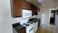 This single-story space includes a kitchen designed for efficient use of its layout. The kitchen is outfitted with rich, dark wood cabinetry that extends to the upper walls, providing generous storage throughout. Dark granite-style countertops contrast with the white appliances, which include an electric coil range, an over-the-range microwave, a dishwasher, and a full-size refrigerator. A double basin sink is positioned along the counter run, accompanied by additional lower cabinetry for added storage. The light-toned flooring in the adjoining area offers a neutral complement to the darker kitchen finishes. The space is practical and well-equipped, with a layout that keeps appliances and work surfaces within easy reach. Natural light filters in, contributing to a bright and functional atmosphere. The overall design emphasizes utility while maintaining a cohesive aesthetic through the consistent use of dark wood tones and coordinating countertop surfaces throughout the kitchen area.