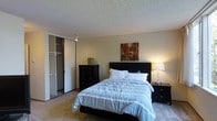 This single-story space includes a bedroom that demonstrates a practical and comfortable layout. The room features plush wall-to-wall carpeting that adds warmth underfoot, complemented by neutral-toned walls that create a calm and inviting atmosphere. A textured popcorn ceiling adds a classic touch overhead.

The bedroom is centered around a queen-sized bed with a dark espresso-finish headboard, flanked by a matching nightstand with a elegant table lamp providing soft, warm illumination. A dark-finish dresser sits nearby, offering generous storage. A piece of wall art in warm earth tones adds a decorative accent above the headboard.

A notable feature of the room is the built-in closet with sliding mirror doors, revealing a well-organized interior with multiple shelving units and hanging rods, maximizing storage potential. The room's layout allows for comfortable movement throughout the space. Overall, the bedroom exudes a cozy, understated ambiance suited for restful living, with functional storage solutions and warm lighting contributing to a welcoming environment.