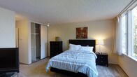 This single-story space includes a bedroom that demonstrates a practical and comfortable layout. The room features plush wall-to-wall carpeting that adds warmth underfoot, complemented by neutral-toned walls that create a calm and inviting atmosphere. A textured popcorn ceiling adds a classic touch overhead.

The bedroom is centered around a queen-sized bed with a dark espresso-finish headboard, flanked by a matching nightstand with a elegant table lamp providing soft, warm illumination. A dark-finish dresser sits nearby, offering generous storage. A piece of wall art in warm earth tones adds a decorative accent above the headboard.

A notable feature of the room is the built-in closet with sliding mirror doors, revealing a well-organized interior with multiple shelving units and hanging rods, maximizing storage potential. The room's layout allows for comfortable movement throughout the space. Overall, the bedroom exudes a cozy, understated ambiance suited for restful living, with functional storage solutions and warm lighting contributing to a welcoming environment.