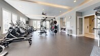 Located at 85 Washington St in Plainville, MA, this fitness center offers a thoughtfully designed, single-floor layout comprising two dedicated exercise rooms and a connecting hallway. The space is generously sized, providing ample room for a wide variety of workout activities without feeling cramped.

The main exercise room impresses with its elegant tray ceiling, recessed lighting, and ceiling fans that maintain comfortable airflow throughout. Large windows invite natural light and views of the outdoors, enhancing the overall ambiance. The durable rubber flooring throughout both rooms provides safety and functionality. This primary space is outfitted with a full power rack with a heavy bag, a cable machine, multiple cardio machines including treadmills, ellipticals, and a recumbent bike, as well as adjustable benches and a wall-mounted television.

The second exercise room is dedicated to free weights and functional training. It features a full dumbbell rack, stability balls, resistance equipment, and adjustable benches. A large wall mirror allows for form checks during workouts, and a ceiling fan keeps the room well-ventilated. Natural light filters in through windows, creating an inviting training environment.

A hallway connects the two rooms and includes storage, a coat rack, and a water station, providing convenient amenities for users. The facility's neutral gray tones and clean, modern finishes contribute to a professional and motivating atmosphere throughout.