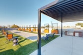 Located at 375 Gartrell St SE in Atlanta's Grant Park neighborhood, this expansive rooftop amenity level offers a thoughtfully designed outdoor experience with sweeping, unobstructed views of the Atlanta skyline. The space is anchored by a central bocce ball court finished in compacted gravel, flanked on both sides by lush artificial turf panels that provide a vibrant, low-maintenance green aesthetic. Warm-toned cedar planter boxes and metal trellis panels are arranged throughout, adding vertical interest and a sense of structure to the open-air layout. A sleek steel pergola with louvered slats defines a shaded lounge area outfitted with modern seating and a gas grill, making it ideal for outdoor gatherings and casual entertaining. Additional seating clusters, bistro-style tables, and accent chairs are distributed across the turf and hardscape areas, accommodating a variety of group sizes. The perimeter features a cable-rail and chain-link safety enclosure that preserves the panoramic views of the Atlanta skyline without obstruction. The surrounding cityscape, including the prominent downtown high-rises, is visible from nearly every vantage point on the rooftop, creating a dramatic urban backdrop. The open design and abundant natural sunlight make this rooftop a standout communal amenity.