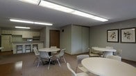 This single-floor space is designed as a functional communal area, combining a dining and lounge setting with an integrated kitchenette. The main room is anchored by multiple round tables paired with lightweight, modern chairs, creating a flexible arrangement well-suited for group meetings, meals, or casual gatherings. The neutral gray walls and carpeted flooring give the space a calm, understated aesthetic throughout.

Overhead, fluorescent light fixtures run across the ceiling, providing consistent and ample illumination throughout the room. A set of large sliding partition panels along one wall offers the potential to divide or reconfigure the space as needed, adding versatility to the layout.

Adjacent to the main area, a compact kitchenette is visible, featuring upper cabinetry, a countertop workspace, and bar-style seating along a raised counter. This functional nook supports food preparation and serving, making the space convenient for communal dining.

A single entry door provides access to the room, and the overall design prioritizes practicality and ease of use. The straightforward, no-frills interior reflects a space intended for high functionality, comfortably accommodating multiple users at once while maintaining an organized and welcoming environment.