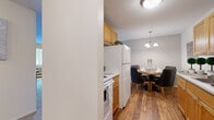 This single-floor residence offers a thoughtfully designed layout that makes efficient use of its space across five distinct rooms. The galley-style kitchen is lined with warm honey-toned wood cabinetry on both sides, providing ample storage, and features hardwood flooring, a double sink, a dishwasher, a white range, and a full-size refrigerator with overhead microwave. A set of louvered closet doors at the end of the kitchen adds additional storage convenience. Adjacent to the kitchen, the dining area is illuminated by an elegant chandelier and showcases the same rich hardwood flooring, creating a seamless and inviting space for meals. The living room is carpeted and filled with natural light streaming through a large picture window with a lush outdoor view, lending a bright and airy ambiance to the space. The bedroom, also carpeted, features a window with a peaceful outdoor tree-lined view, a ceiling fan, and generous room for furnishings. The bathroom is finished in crisp white tones, featuring a wide vanity with ample counter space, a single sink, and a shower-tub combination. Throughout the unit, a textured ceiling and neutral wall palette create a clean, cohesive look.