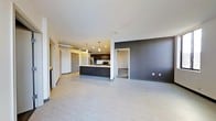 This single-floor space showcases a thoughtfully designed open-concept layout that seamlessly integrates the living area and kitchen. The kitchen is a standout feature, centered around a dark-finished island with pendant lighting overhead, complemented by stainless steel appliances including a refrigerator, microwave, and range. Dark cabinetry adds a bold contrast against the neutral-toned flooring that runs throughout the space. An exposed metallic ventilation duct runs along the ceiling, lending an industrial character to the overall aesthetic. A dramatic dark accent wall in the main living area creates a striking focal point and adds depth to the open floor plan. The light gray flooring provides a clean, cohesive foundation that ties the space together. Additional rooms are accessible through doorways visible from the main living area, offering functional separation while maintaining an open feel. Recessed lighting throughout ensures consistent illumination, while the smooth white ceilings enhance the sense of openness. The overall design balances modern minimalism with industrial accents, resulting in a stylish and functional living environment.