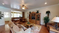 This single-floor residence offers a welcoming and cohesive interior defined by warm wood tones, hardwood flooring, and classic crown molding throughout. The open living area serves as the heart of the home, centered around a substantial wooden entertainment unit and accented by a ceiling fan with recessed lighting above. A floral area rug anchors comfortable seating, while large windows allow natural light to fill the space and offer views of the surrounding greenery. The living area flows seamlessly into a dedicated dining space, which features a round table with seating for multiple guests, positioned near a pass-through opening to the kitchen. The kitchen is functional and well-appointed, featuring warm wood cabinetry, stainless steel appliances, a double sink, and a row of windows that bring in abundant natural light. Countertops provide ample prep space, complemented by a skylight-style ceiling fixture overhead. Throughout the interior, framed artwork, potted plants, and tasteful décor contribute to a cohesive, comfortable ambiance. Outside, residents enjoy access to a generously sized community pool with a spa, shaded by mature trees and outfitted with lounge seating and red umbrella-covered tables. A charming white gazebo nestled amid lush lawns and manicured landscaping adds a tranquil retreat within the community grounds.