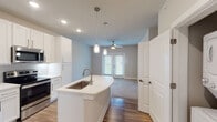 This single-story residence in Panama City Beach, Florida, offers a well-designed open-concept layout that maximizes its comfortable footprint. The kitchen serves as the centerpiece of the living area, featuring white shaker-style cabinetry extending to the ceiling, light quartz countertops, a subway tile backsplash, and a full suite of stainless steel appliances including a refrigerator, electric range, and built-in microwave. A curved kitchen island with an undermount sink and pendant lighting provides both additional workspace and a natural division between the kitchen and living area. Wood-style flooring runs throughout the main living spaces, complementing the neutral gray and white color palette. The living area receives natural light through French doors that lead to the exterior, with a ceiling fan adding functional comfort. The bedroom is carpeted for warmth, with a large window allowing ample natural light. The bathroom features a single vanity with quartz countertops, an undermount sink, framed mirror, and a glass-enclosed shower, with open shelving providing convenient storage. The overall design presents a clean, modern aesthetic suitable for Florida coastal living.
