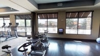 This fitness facility in Flower Mound is thoughtfully designed to support a comprehensive workout experience across several distinct zones. The cycling studio is a standout feature, accommodating multiple stationary bikes on rich, dark hardwood flooring beneath tray ceilings with contrasting gray accents. Large windows fitted with roller shades line the walls, flooding the space with natural light while offering outdoor views of a pool area and surrounding landscape. A mounted digital display panel provides interactive functionality for cyclists.

A floor-to-ceiling glass partition separates the cycling area from the main strength and cardio floor, which houses cable machines, treadmills, and additional equipment on lighter hardwood flooring. This open, well-lit zone benefits from recessed lighting and a clean, modern aesthetic.

A dedicated yoga and media room offers a quieter retreat, featuring a large wall-mounted projection screen flanked by mounted speakers, making it ideal for guided classes. Dark hardwood floors, a workout mat, and stability balls complete the space.

Dark-stained interior doors with bold paneling details provide access to additional rooms, maintaining a cohesive, contemporary design throughout the facility.