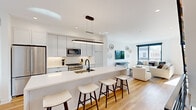 This contemporary two-floor residence showcases a thoughtfully designed open-concept layout with a clean, modern aesthetic throughout. The main living area seamlessly connects the kitchen, dining, and living spaces, all unified by warm light hardwood flooring and crisp white walls that maximize the sense of brightness and openness.

The kitchen is a standout feature, offering a generous island with a quartz countertop, bar seating, matte black fixtures, stainless steel appliances including a gas range and built-in microwave, and sleek flat-panel cabinetry extending to the ceiling. A sculptural oval pendant light anchors the space above. The adjoining dining area benefits from a large sliding glass door that opens to a balcony with urban views, while the living room features oversized windows that flood the space with natural light.

The bedrooms are serene and well-proportioned, each with floor-to-ceiling windows framing neighborhood views. The bathroom is finished with large-format stone-look tile, a glass-enclosed walk-in shower, a floating vanity with dual sinks, and matte black hardware throughout. An additional room with direct outdoor access provides flexible living options. The overall ambiance is polished, light-filled, and contemporary.