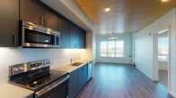 Located at 1090 NE 91st Ave in Hillsboro, Oregon, this thoughtfully designed single-floor residence combines modern finishes with functional living spaces. The kitchen is a standout feature, showcasing sleek dark navy cabinetry, light quartz countertops, a white subway tile backsplash, and a full suite of stainless steel appliances including a refrigerator, electric range, over-the-range microwave, and dishwasher. A distinctive warm wood-plank ceiling extends from the kitchen into the open living area, adding a natural, inviting contrast to the clean white walls and dark hardwood-style flooring beneath. The living area is generously proportioned, illuminated by a ceiling fan with integrated lighting and a large window that frames outdoor views and draws in abundant natural light. The bedroom continues the warm aesthetic with carpeted flooring, a ceiling fan, wood-plank ceiling detail, and a bright window overlooking the surrounding landscape. The bathroom is well-appointed with a soaking tub and shower combination, built-in wall shelving, a dark-toned vanity with an undermount sink, and hardwood-style flooring. The overall design balances contemporary style with comfortable, practical living throughout.