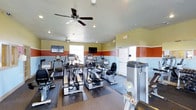 This fitness center is a fully outfitted workout facility designed to meet a wide range of exercise needs. The space is thoughtfully arranged to maximize usability, featuring a lineup of modern cardio equipment including treadmills, elliptical trainers, and stationary bikes from recognized brands such as TRUE Fitness. Additional strength and conditioning equipment is also present, rounding out the facility's offerings.

The interior is bright and energizing, with large windows allowing ample natural light to fill the space. A bold color scheme — featuring bands of terracotta orange, soft yellow, and light blue along the walls — gives the room a dynamic, motivating atmosphere. Recessed lighting and dual ceiling fans with integrated lighting ensure comfortable illumination and air circulation throughout.

Multiple wall-mounted flat-screen televisions are positioned at convenient viewing angles, allowing users to stay entertained during workouts. The flooring is finished with durable rubber matting beneath the equipment, providing safety and stability. The single-floor layout ensures everything is easily accessible, making this a functional and welcoming environment for fitness enthusiasts of all levels.