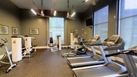 This fitness center is thoughtfully designed to support a well-rounded workout experience. The space is outfitted with a selection of modern cardio equipment, including treadmills, a stationary bike, and an elliptical machine, alongside dedicated strength training stations that cater to a variety of exercise routines. The flooring features durable rubber gym matting, providing both safety and comfort during workouts. The room is illuminated by a combination of sleek overhead linear lighting and ample natural light streaming through large windows, creating a bright and energizing atmosphere. Ceiling fans are suspended from above, ensuring good air circulation throughout the space. A wall-mounted flat-screen television adds an entertainment option for those looking to stay engaged during cardio sessions. The walls are painted in a deep charcoal tone, lending the space a focused and purposeful ambiance. Motivational signage bearing words such as "Believe" and "Discipline" adorns the walls, reinforcing an environment of determination and drive. The overall design is clean, functional, and modern, making this fitness center a practical and inviting space for daily exercise.