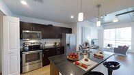 This single-floor residence showcases a thoughtfully designed open-concept layout that maximizes both functionality and style. The kitchen serves as the centerpiece of the space, featuring rich dark cabinetry that contrasts beautifully with crisp white subway tile backsplash. A large kitchen island with dark granite countertops provides generous prep space and casual seating, illuminated by modern pendant lighting overhead. Stainless steel appliances, including a French door refrigerator with ice and water dispenser, a range, and an over-the-range microwave, complete the contemporary culinary suite. The sink is elegantly integrated into the island, adding to the kitchen's practical appeal. Light hardwood flooring extends throughout, creating a warm and cohesive flow from the kitchen into the dining and living areas. The open sightlines allow natural light to permeate the entire space, enhancing its airy and welcoming ambiance. Track lighting and recessed fixtures ensure well-distributed illumination throughout. The seamless transition between the kitchen, dining, and living zones makes this residence ideal for both everyday living and entertaining.