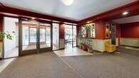 Located at 17000 Chemin Sainte-Marie in Kirkland, Quebec, this shared common area encompasses a spacious lobby and an adjoining fitness room. The lobby makes a strong first impression with its bold red accent walls contrasted against cream lower paneling, recessed lighting, and a combination of tile and carpet flooring that defines distinct zones within the space. Lush potted plants add a welcoming, organic touch to the entrance area, while a framed mirror and a wooden bench seat provide functional, decorative elements near the elevator. A bank of individual mailboxes is conveniently integrated into the lobby wall, and a stainless steel elevator provides easy vertical access throughout the building. Informational boards and secured entry doors complete the practical layout of the entrance zone. Adjacent to the lobby, the fitness room offers a well-rounded selection of exercise equipment, including a multi-station cable machine, a treadmill, and a recumbent bike, all set on warm tile flooring. Painted in soft beige and blue tones, the gym benefits from natural light streaming through a large exterior-facing window. A mirrored wall enhances the sense of space, and a glass partition provides a visual connection back to the lobby. Together, these areas offer residents a functional and welcoming communal environment.