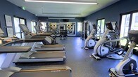 This fitness center offers a comprehensive workout environment designed to accommodate a wide range of exercise needs. The space is finished with deep navy-blue walls that lend a sleek, modern aesthetic, complemented by durable rubber flooring that provides both safety and stability underfoot.

The cardio area is outfitted with a row of Matrix treadmills and elliptical trainers, positioned to take advantage of natural light streaming through large windows. A full-length mirror along one wall visually expands the space and allows users to monitor their form during workouts.

The strength training zone features a multi-functional cable machine at its centerpiece, ideal for a wide variety of resistance exercises. An adjustable weight bench and a set of medicine balls round out the free-weight area, supporting both functional and traditional strength training routines.

Overhead, recessed linear lighting ensures consistent illumination throughout the space. The windows on multiple walls bring in ample natural light and offer views of the surrounding exterior, enhancing the overall atmosphere. Framed artwork adds a subtle decorative touch, balancing the functional nature of the facility with a polished, inviting ambiance.