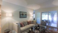 This single-story space centers around a thoughtfully designed living area that balances comfort and style. The room features vaulted ceilings that lend an open, airy feel, with soft, light-toned walls creating a calm and inviting atmosphere. A cream-colored sofa adorned with patterned and warm terracotta accent pillows serves as the focal point, complemented by a matching upholstered armchair in a coordinating geometric print. A glass-top coffee table anchors the seating arrangement, adding a contemporary touch. A landscape painting mounted on the wall introduces a pop of color and visual interest. Natural light flows generously into the space through a sliding glass door, which provides direct access to an outdoor area, offering views of lush greenery beyond. A floor lamp positioned near the window supplements the ambient lighting, creating a warm glow throughout the room. The overall ambiance is relaxed yet polished, with cohesive décor choices that give the space a move-in-ready, welcoming character suitable for both everyday living and entertaining.