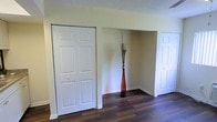 This single-floor space presents a straightforward and functional layout with a cohesive interior design. The flooring throughout is a rich, dark hardwood that adds warmth and a sense of continuity across the open areas. Walls are finished in a soft, neutral tone that complements the overall clean aesthetic of the space.

The main open area is moderately sized, providing enough room for versatile use. A textured ceiling adds subtle visual interest overhead. The space features multiple storage solutions, including classic six-panel bifold and standard panel doors that open to closets, offering ample storage capacity. A recessed nook between two closets provides an additional architectural detail, creating a natural display or storage alcove.

Toward one end of the space, a partially visible adjoining area suggests a secondary room or utility zone, separated by a partial wall partition. A window allows natural light to filter into this section. White trim and baseboards frame the walls throughout, providing a crisp contrast against both the flooring and wall color.

Overall, the space maintains a neutral, move-in ready character with practical storage, durable flooring, and a simple, uncluttered design that allows for flexible arrangement and use.
