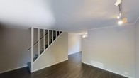 This multi-level space spans three floors, offering a versatile layout designed to maximize living areas across each level. The main living area showcases dark hardwood flooring that contrasts cleanly with the bright white walls and ceiling, creating a crisp, neutral aesthetic throughout. A staircase with white-painted railings and dark treads serves as a functional architectural element, connecting the floors while adding visual structure to the open living space. A ceiling fan is installed overhead, providing both practical air circulation and a subtle design touch. White baseboards frame the perimeter of the room, complementing the overall clean and understated interior palette. The open floor plan of the main level allows for flexible furniture arrangement and a sense of spaciousness. Recessed floor vents suggest a central HVAC system integrated throughout the space. The overall ambiance is clean and move-in ready, with neutral finishes that provide a blank canvas adaptable to a variety of interior styles. The three-floor configuration offers ample separation between living, sleeping, and utility spaces.
