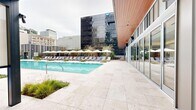 This two-level outdoor amenity space is a masterfully designed urban retreat set against a dramatic downtown skyline. At its centerpiece sits a generously sized lap pool finished with travertine tile surrounds, flanked by rows of lounge chairs and white shade umbrellas that create a resort-like atmosphere amid the city environment.

The pool deck is thoughtfully landscaped with ornamental grasses and sculpted plantings, softening the contemporary hardscape. Adjacent to the pool, a covered outdoor kitchen area features a built-in stainless steel grill set into warm wood cabinetry, paired with an outdoor refrigerator and ample counter space for food preparation and entertaining.

A covered lounge pavilion with a rich wood-slatted ceiling provides a shaded gathering area complete with a long communal bar-height table surrounded by woven rattan counter stools. An outdoor projector screen mounted nearby adds an entertainment dimension to the space. Dual outdoor showers backed by a horizontal wood slat privacy wall offer convenient poolside functionality. Floor-to-ceiling glass walls on the adjacent building connect the outdoor space seamlessly to the interior, maximizing natural light and city views throughout.