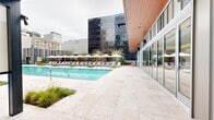 This two-level outdoor amenity space is a masterfully designed urban retreat set against a dramatic downtown skyline. At its centerpiece sits a generously sized lap pool finished with travertine tile surrounds, flanked by rows of lounge chairs and white shade umbrellas that create a resort-like atmosphere amid the city environment.

The pool deck is thoughtfully landscaped with ornamental grasses and sculpted plantings, softening the contemporary hardscape. Adjacent to the pool, a covered outdoor kitchen area features a built-in stainless steel grill set into warm wood cabinetry, paired with an outdoor refrigerator and ample counter space for food preparation and entertaining.

A covered lounge pavilion with a rich wood-slatted ceiling provides a shaded gathering area complete with a long communal bar-height table surrounded by woven rattan counter stools. An outdoor projector screen mounted nearby adds an entertainment dimension to the space. Dual outdoor showers backed by a horizontal wood slat privacy wall offer convenient poolside functionality. Floor-to-ceiling glass walls on the adjacent building connect the outdoor space seamlessly to the interior, maximizing natural light and city views throughout.