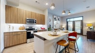 This single-floor residence showcases a thoughtfully designed open-concept layout that seamlessly connects the kitchen, dining, and living areas. The kitchen is a standout feature, equipped with flat-front wood-tone cabinetry extending to the ceiling for ample storage, stainless steel appliances including a range, microwave, refrigerator, and dishwasher, and a quartz-topped island with seating that doubles as a casual dining space. Pendant lighting above the island adds a contemporary touch, while light hardwood-style flooring runs throughout the main living areas.

The living room is bright and welcoming, anchored by floor-to-ceiling doors that open to an outdoor area and allow generous natural light to filter in. The space comfortably accommodates a seating arrangement and benefits from a ceiling fan overhead.

Both bedrooms are carpeted for comfort, feature large windows with outdoor views, and offer a calm, neutral palette. The full bathroom includes a wood-tone vanity with quartz countertops, a tub-shower combination with white tile surround, and light vinyl flooring, maintaining the cohesive modern aesthetic found throughout the residence.