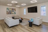Located at 706 Huffman Mill Rd in Burlington, NC, this single-floor commercial space offers a polished and contemporary environment designed to welcome visitors and support professional activity. The main lounge area serves as the heart of the space, characterized by crisp white walls, warm wood-look flooring, and a drop ceiling outfitted with distinctive diamond-shaped LED light fixtures that provide bright, even illumination throughout. The seating arrangement is thoughtfully composed, featuring neutral-toned sofas and accent chairs arranged around sleek, dark side tables and a textured area rug, creating a comfortable yet refined atmosphere. A wall-mounted flat-screen television anchors one side of the room, flanked by windows with blinds that allow natural light to filter in while maintaining privacy. Decorative framed artwork adorns the walls, adding visual interest and a touch of sophistication. French-style interior doors with glass panes connect the lounge to adjacent areas, including what appears to be a fitness or equipment room visible in the background, suggesting the space supports a range of functional uses. The overall design aesthetic is clean, modern, and professionally appointed, making it well-suited for client-facing or wellness-oriented business operations.